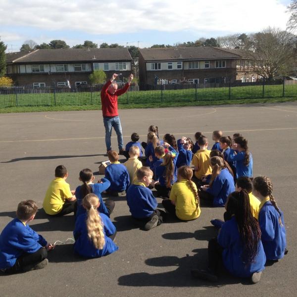 Man teaching about solar power to group of children sitting on ground in school playground