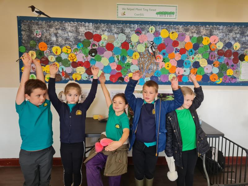 5 primary school kids waving in front of collage of pledges to celebrate planting the Tiny Forest Caerphilly