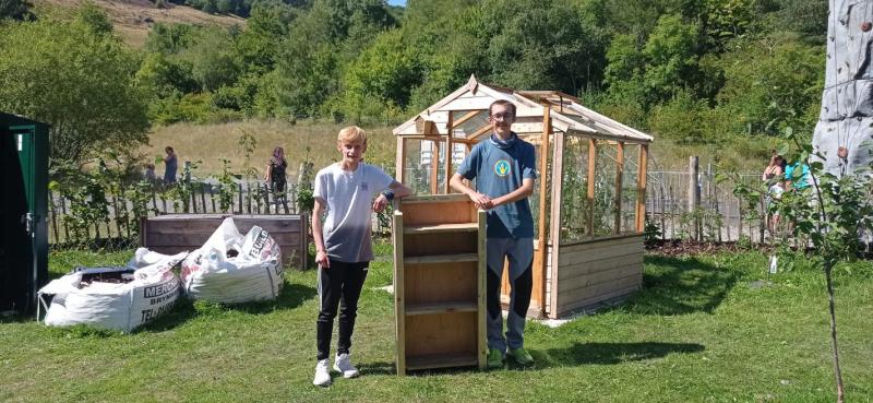 Man and boy holding up seed library shelves, garden and greenhouse in background