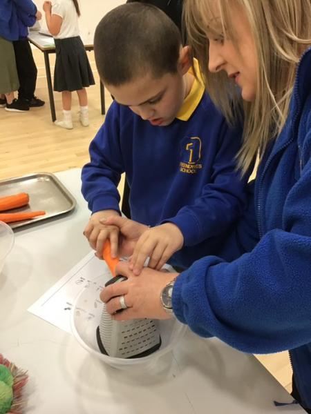 Close up of blond woman helping small schoolboy to grate carrots on classroom table