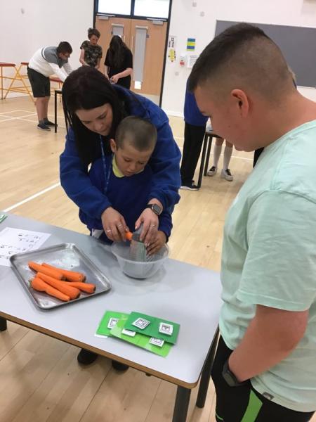 Woman helping small child to grate carrots in classroom situation, older boy looking on