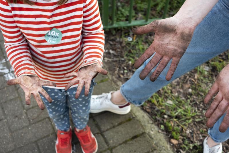 Child wearing a red and white striped t-shirt shows grubby hands to adult whose hands are also covered in soil, at concrete-smashing and planting event in hackney E5