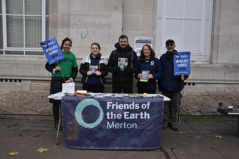 5 people standing behind a street stall covered in a Merton FOE banner holding placards about insulating homes. 
