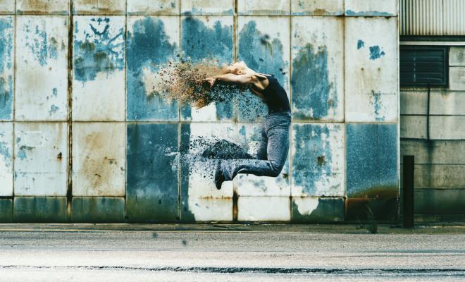 Female dancer jumps high up in the air, her body dissolves and fragments fly off her head, back and legs. In the background is an old industrial building.