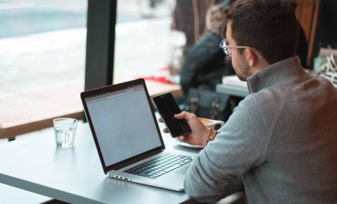 Man sitting at desk with open laptop, checking his mobile phone