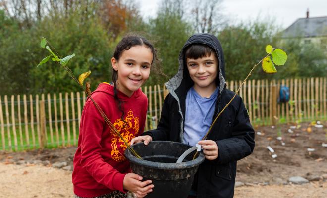 Two children holding a sapling in a bucket on Tiny Forest Caerphilly site on planting day