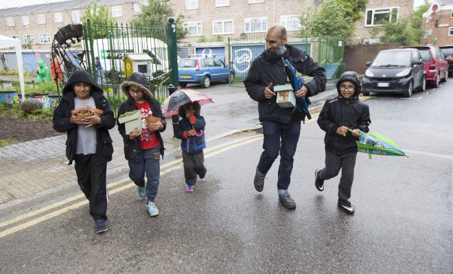 Farther and 4 children leaving community garden and crossing road in a line, carrying plants from 10xG launch