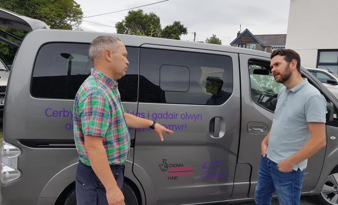 2 men in conversation beside electric vehicle, partly obsured Welsh text on side of car.