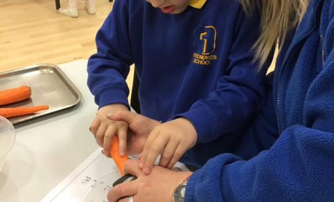 Close up of blond woman helping small schoolboy to grate carrots on classroom table