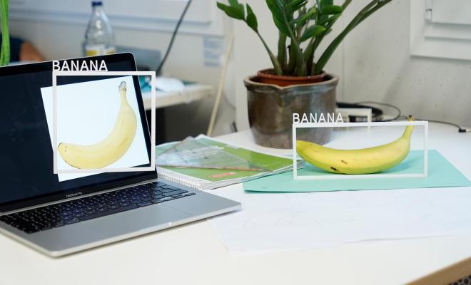 A banana on a table and an image of a banana on a laptop on the same table. Each of the two bananas has a white frame around it with the word ‚Banana‘ stuck on top of it