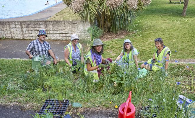POstcode Gardener Lizz Dobinson surrounded by volunteers working on a bank covered in plants looking up at camera, paddling pool in background