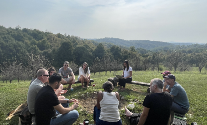 People dressed imnformally seated in a circle in the sunshine, country landscape in background