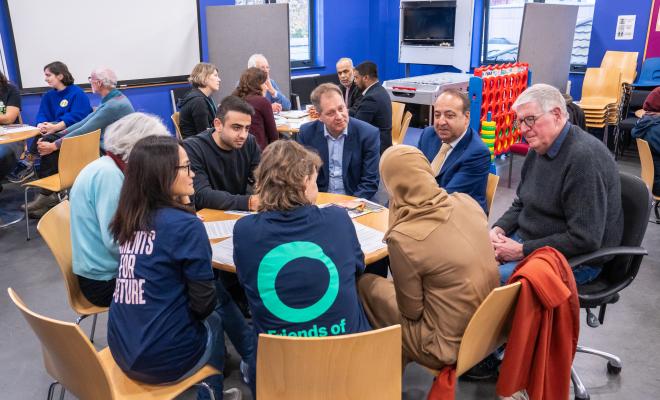 3 young women, one in FOE t-shirt, sit round table in a meeting with men in suits